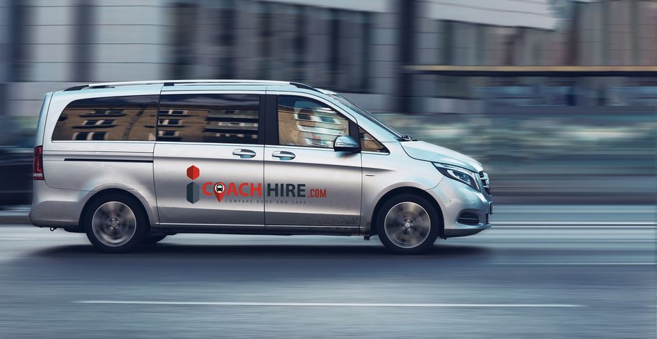 A silver minibus with icoachhire.com written on the side, in motion in an urban area